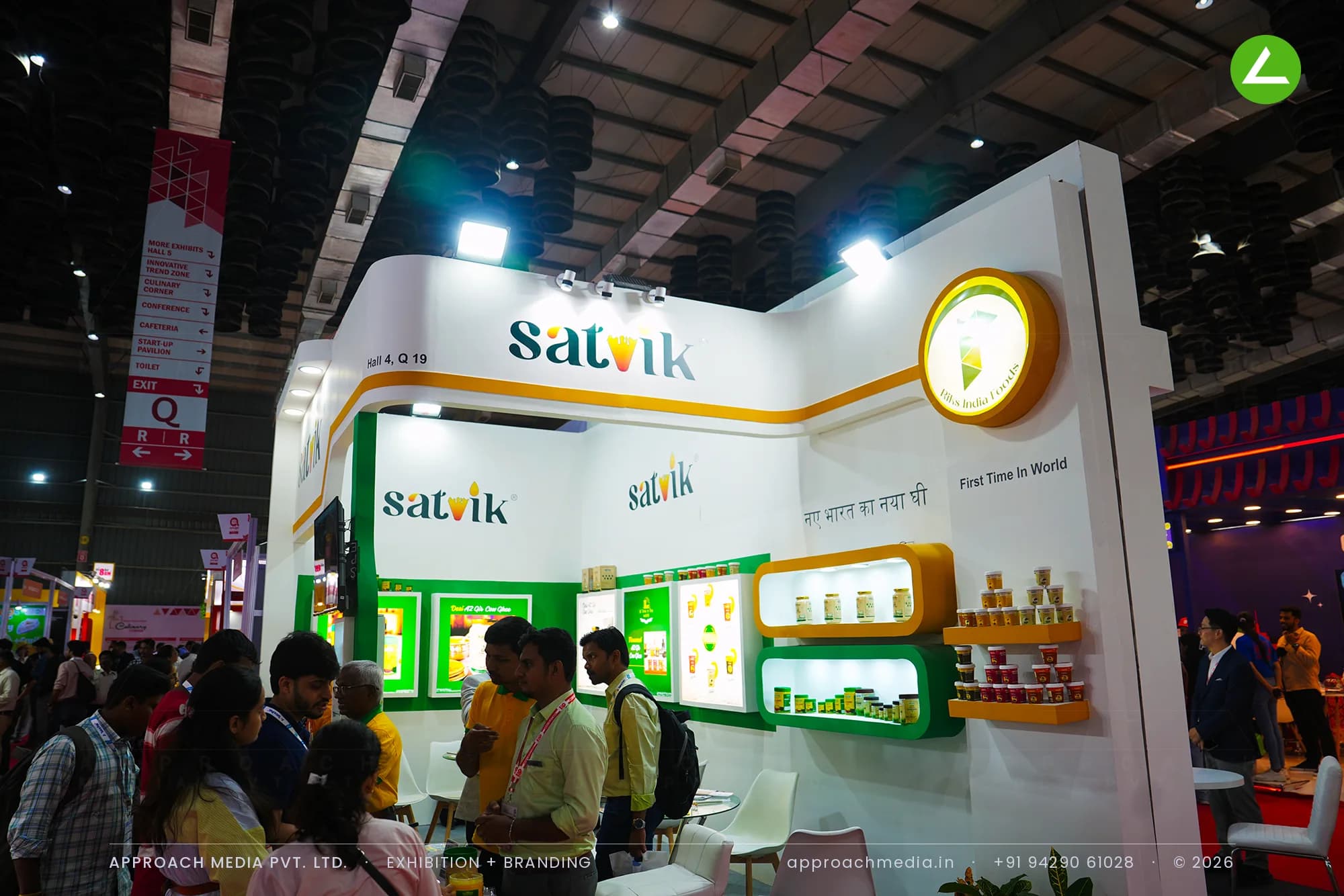 A white fascia wraps Satvik's corner with an orange ribbon and a lit disc sign, above orange niches holding jars of ghee.