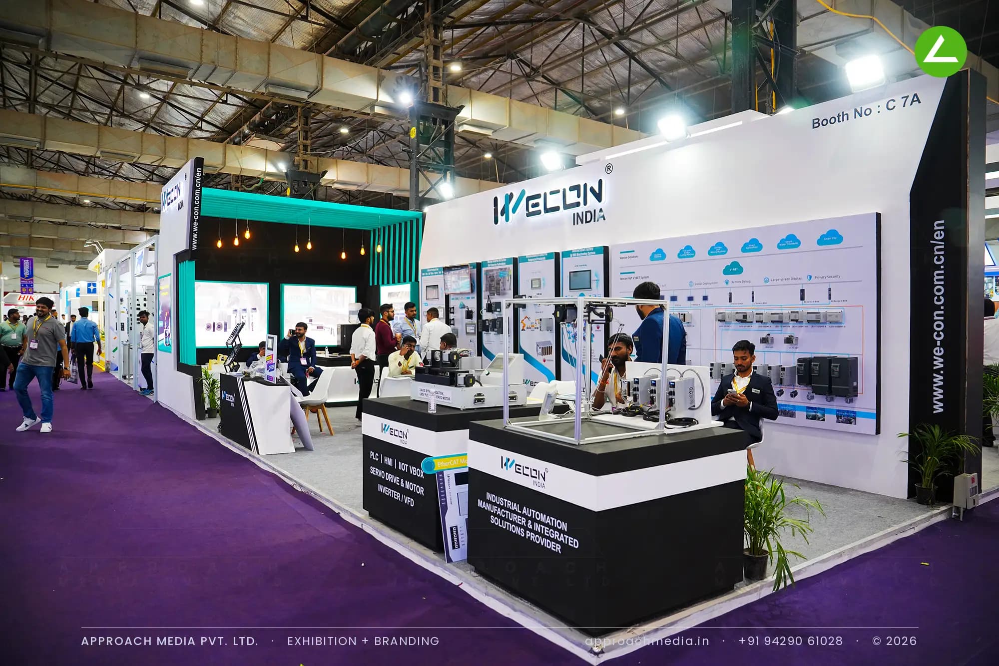 White demo counters with black tops stand in front of Wecon India's printed controller wall, below a teal slatted canopy.